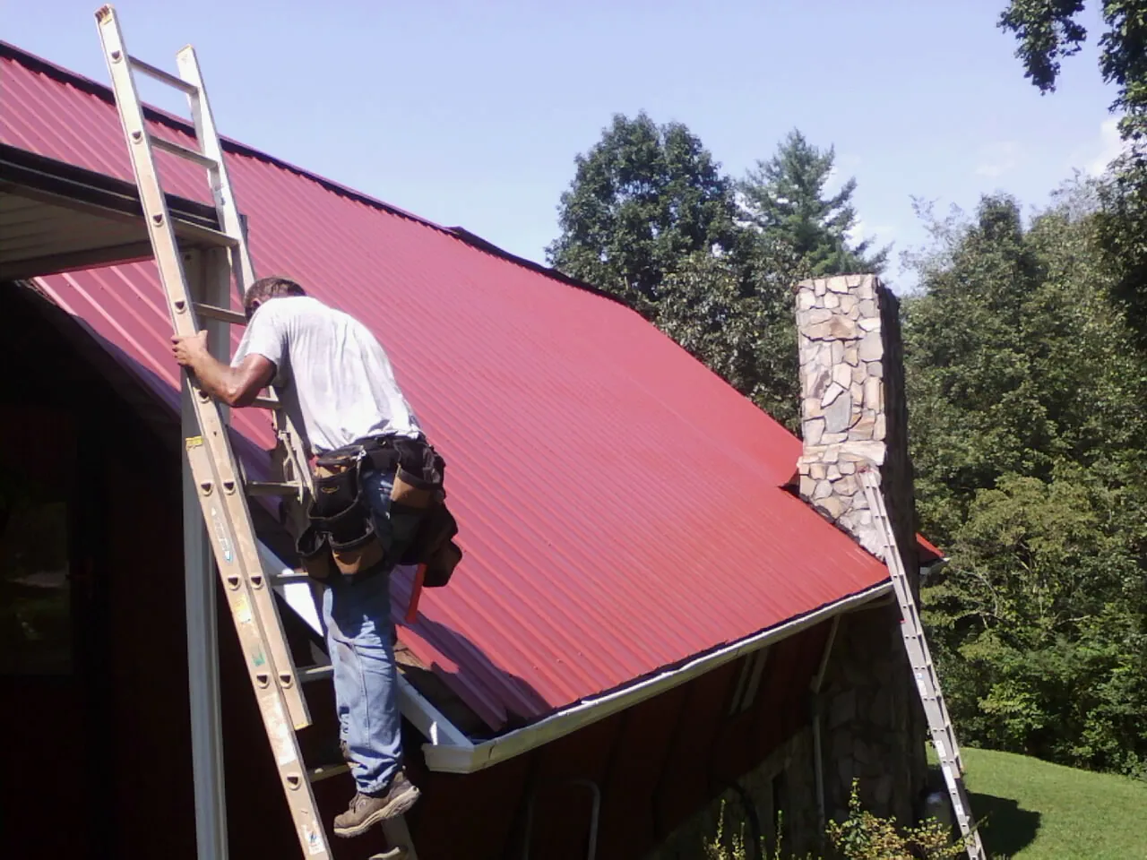 Roofer on a ladder beside a red metal roof and stone chimney — Soffit & Fascia Repair by Roofing Brothers in Hunter